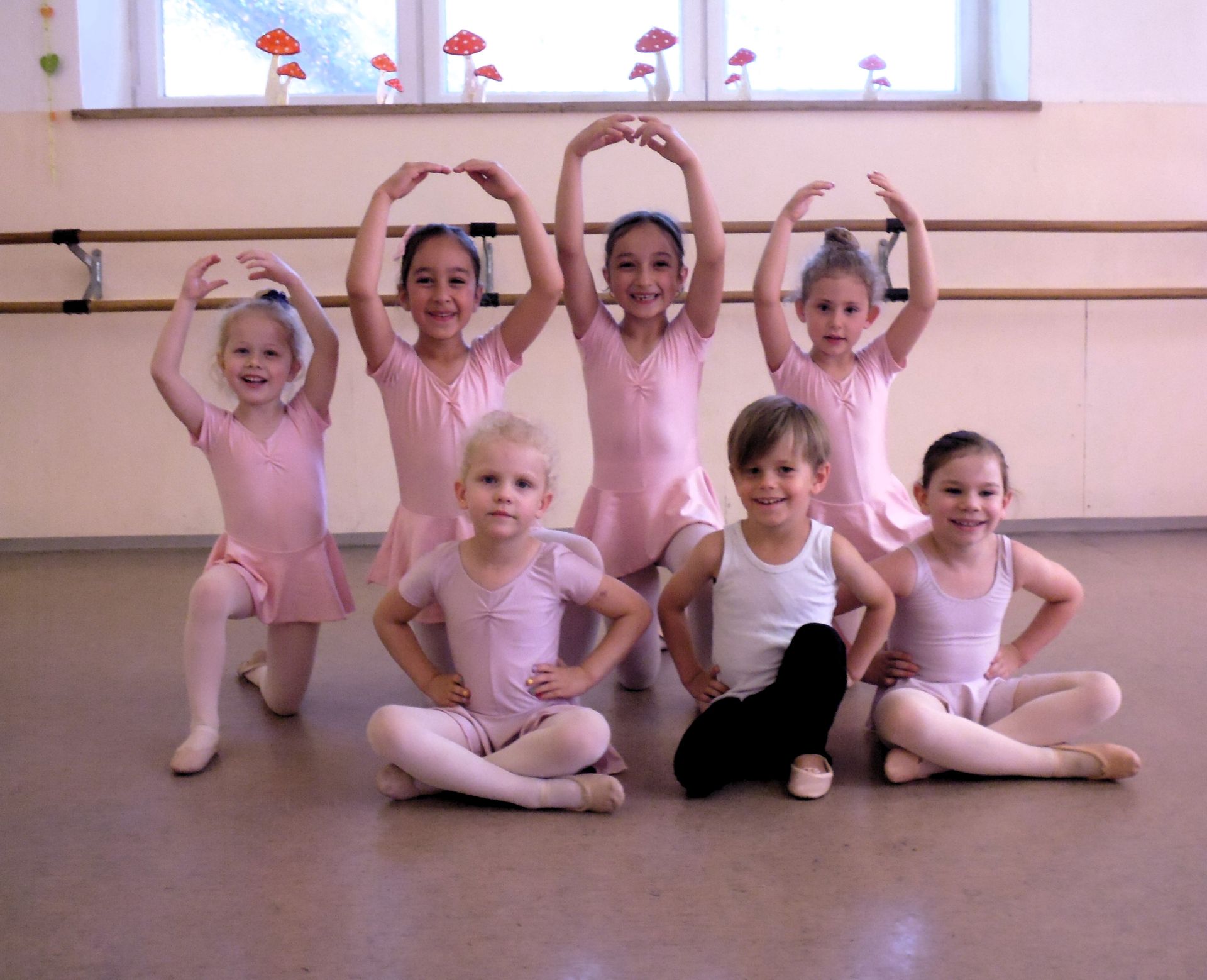 Kinder in rosa Ballettkostümen posieren in einem Tanzstudio mit einer hölzernen Ballettstange und Fenstern.