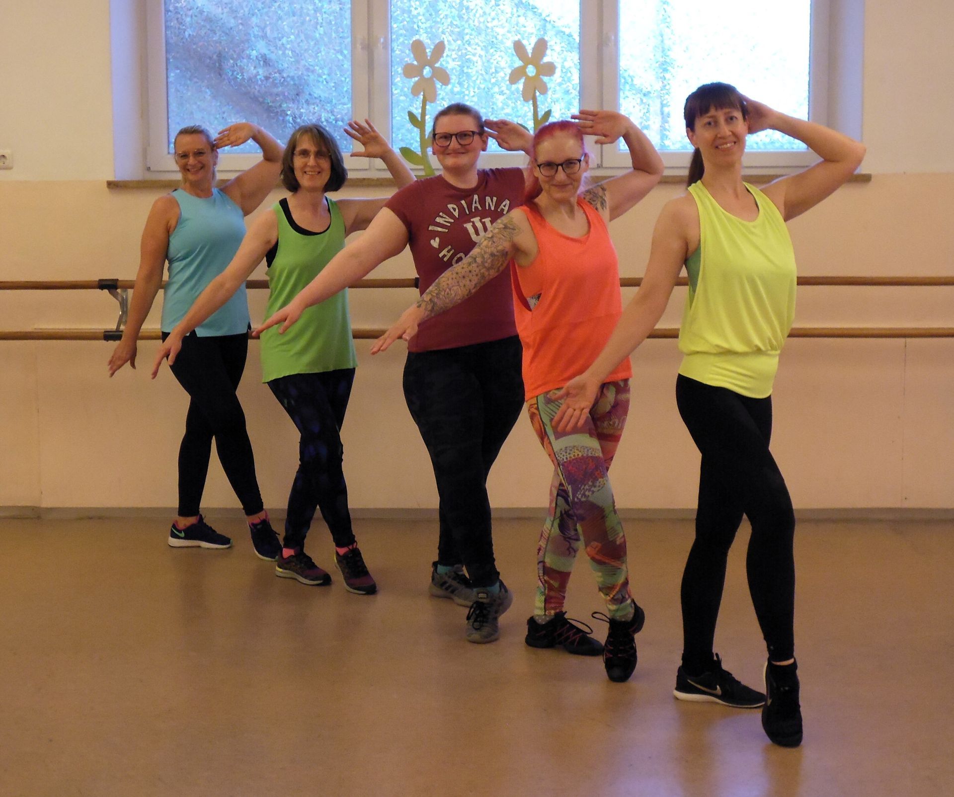 Menschen tanzen in einem hell erleuchteten Fitnessstudio, angeführt von einer Frau in einem pinkfarbenen Tanktop.
