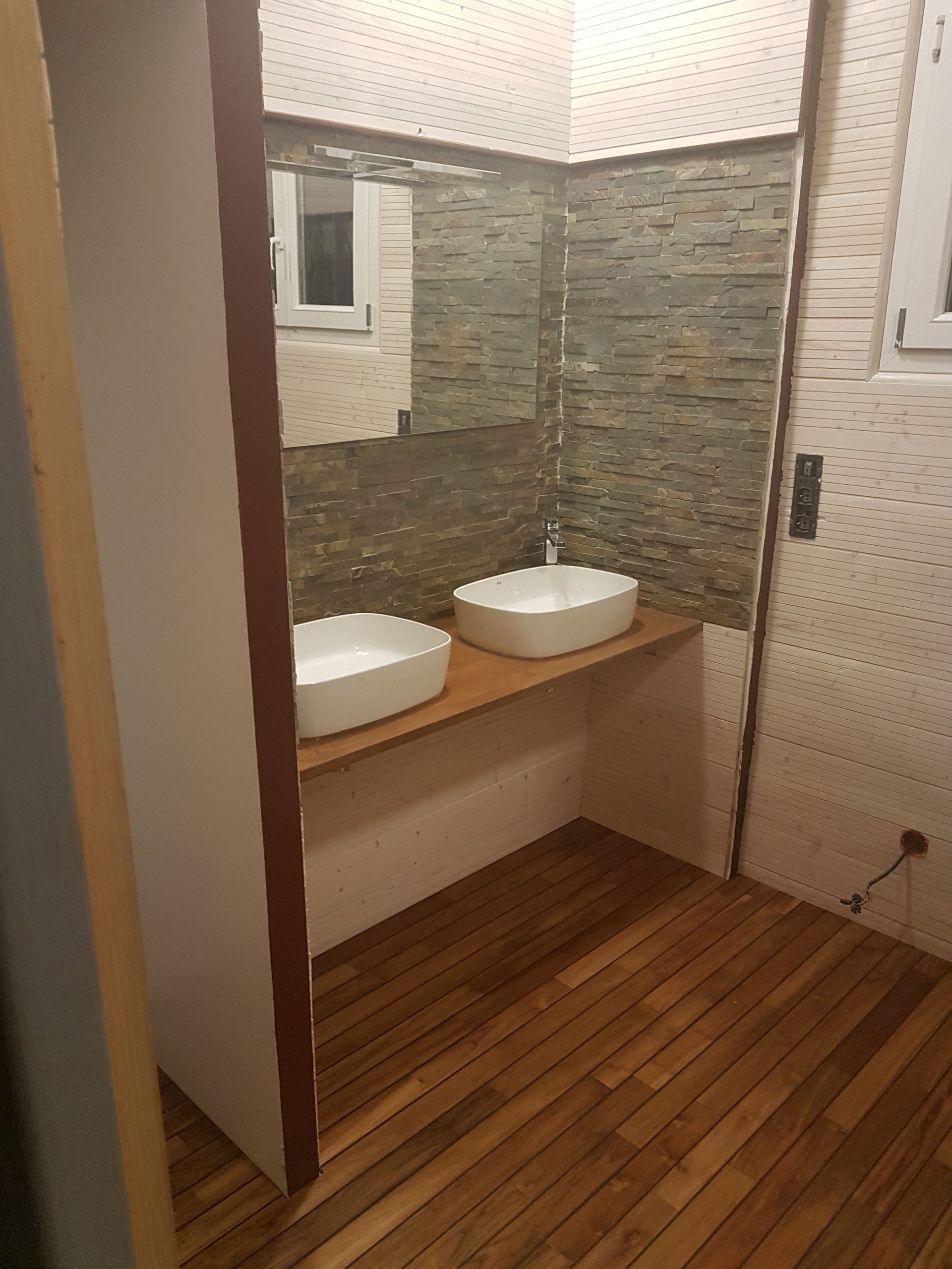 Meuble-lavabo moderne avec deux vasques blanches, comptoir en bois, mur carrelé en pierre et parquet à rayures.