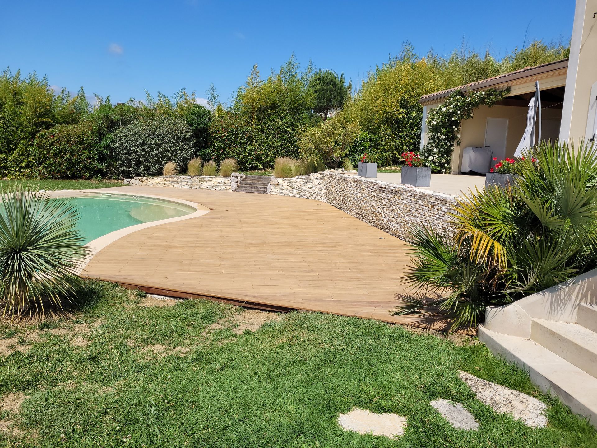 Terrasse en bois incurvée au bord d'une piscine, avec un jardin luxuriant et un patio sous un ciel bleu azur.