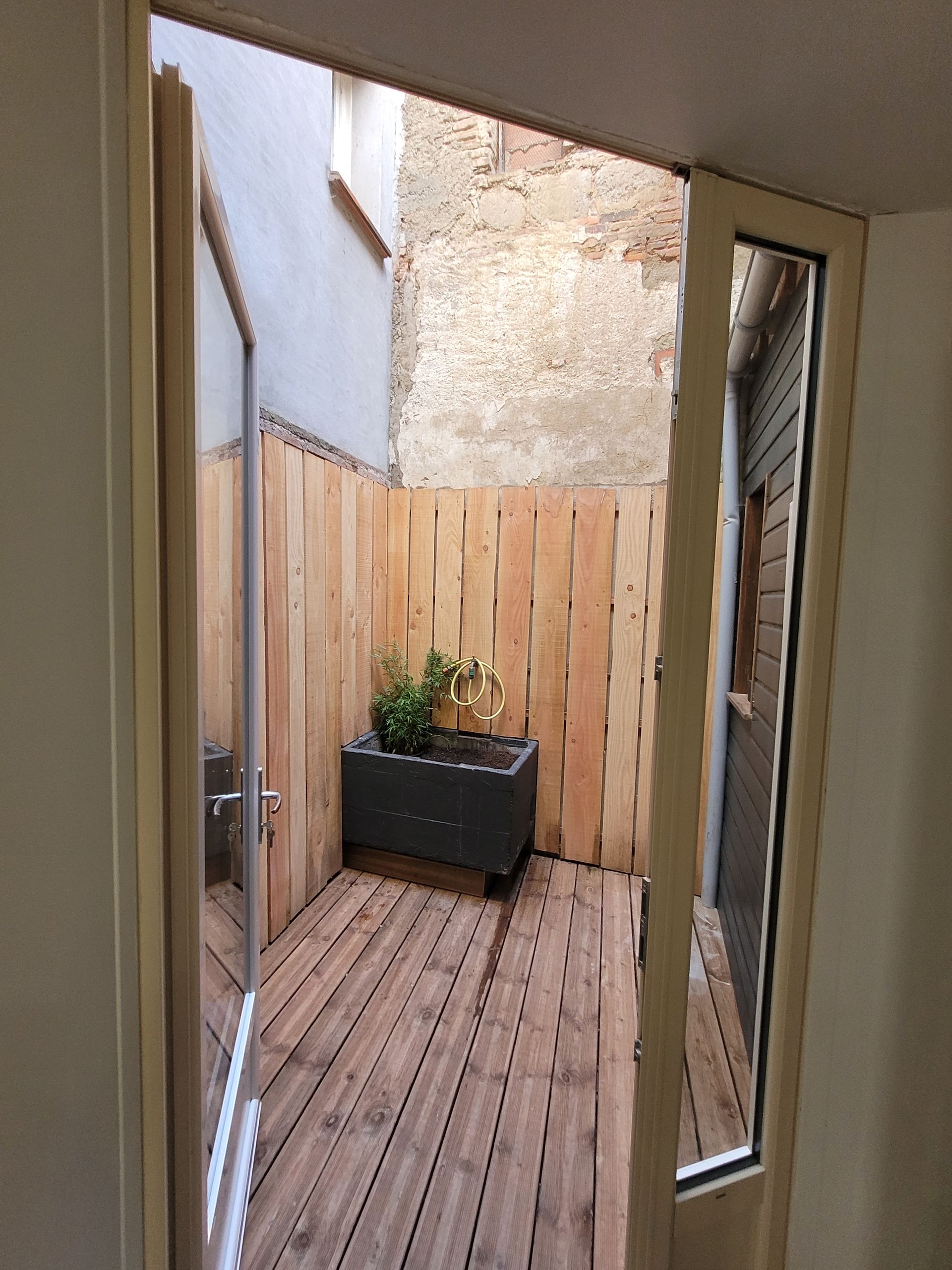 Étroite terrasse en bois avec plante en pot, entourée de clôtures et de murs en pierre, visible à travers des portes vitrées ouvertes