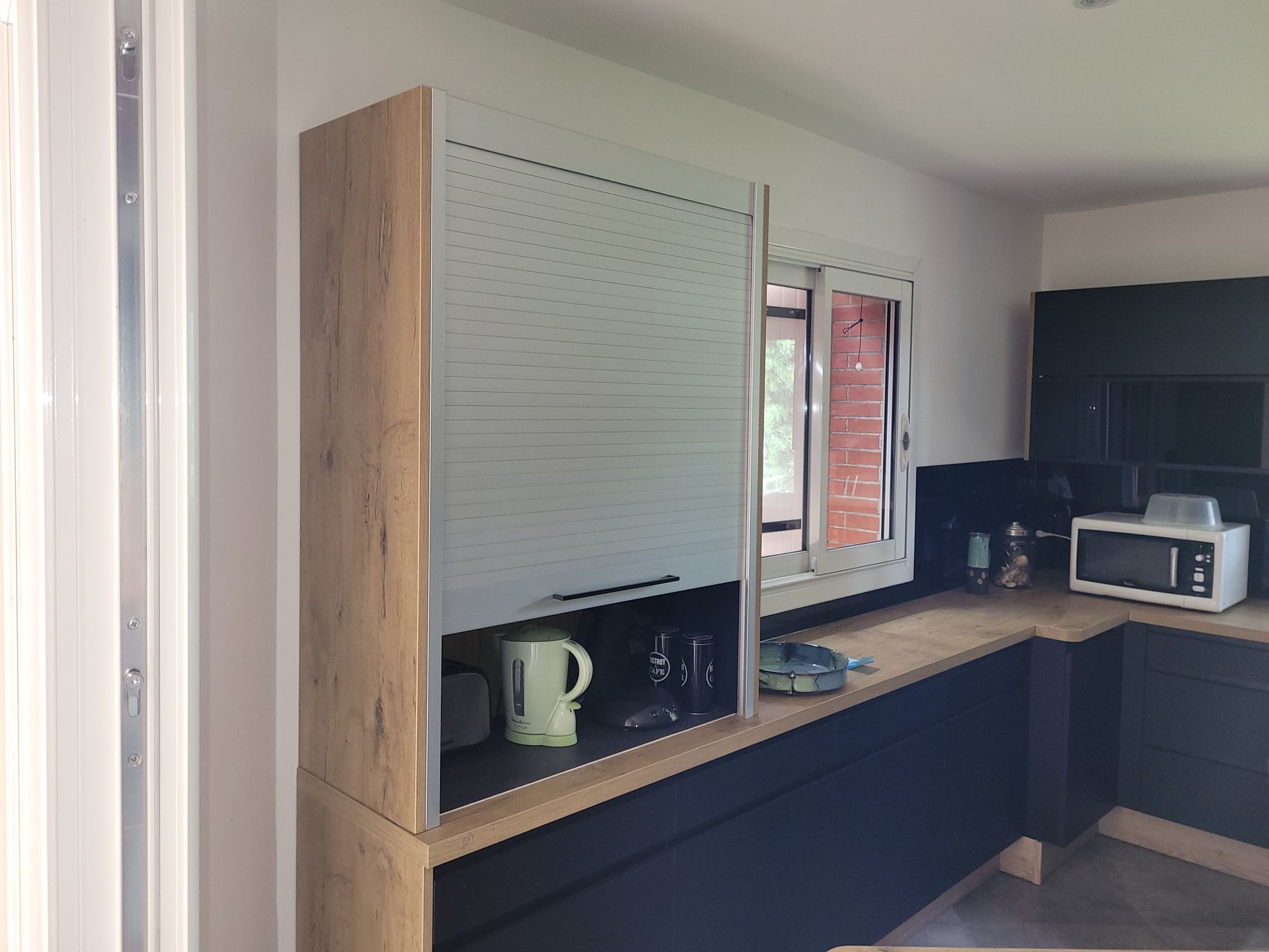 Cuisine moderne avec placards en bois, plans de travail noirs, micro-ondes, bouilloire et stores.