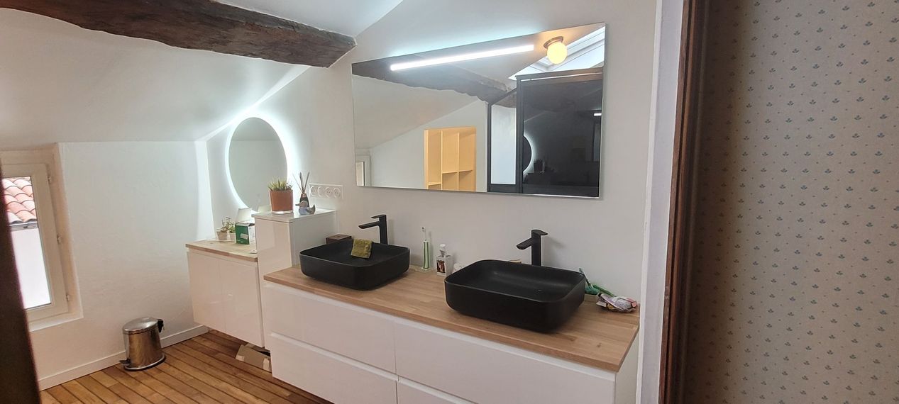 Meuble-lavabo moderne avec double vasque, miroir rond éclairé et comptoir en bois sous un plafond incliné