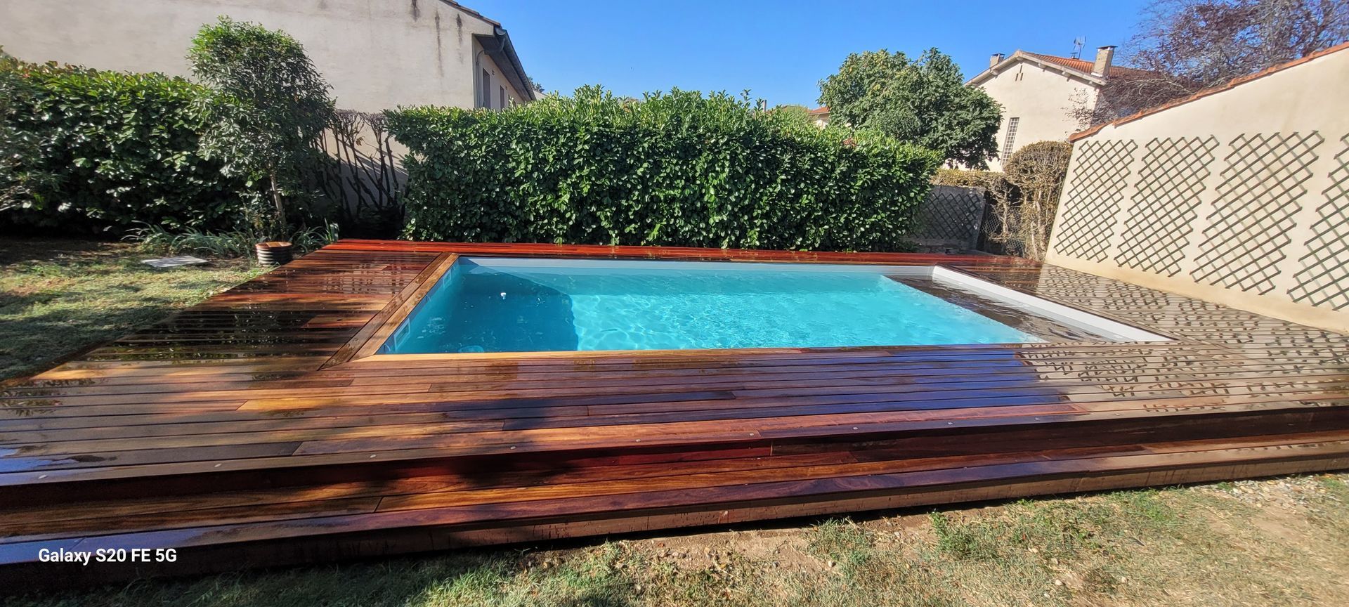 Piscine dans la cour arrière avec terrasse en bois, arbustes verts et maisons voisines sous un ciel dégagé