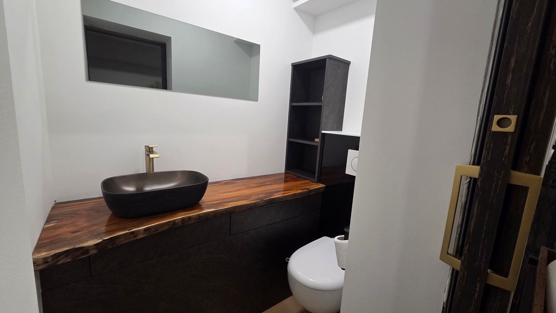 Salle de bains moderne avec comptoir en bois, vasque noire, miroir et toilettes blanches.