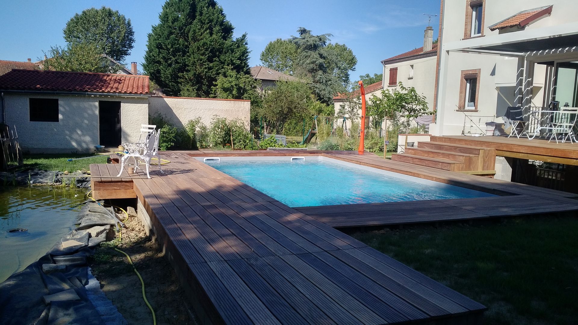 Piscine dans le jardin avec terrasse en bois, chaises longues et maison blanche baignée de soleil