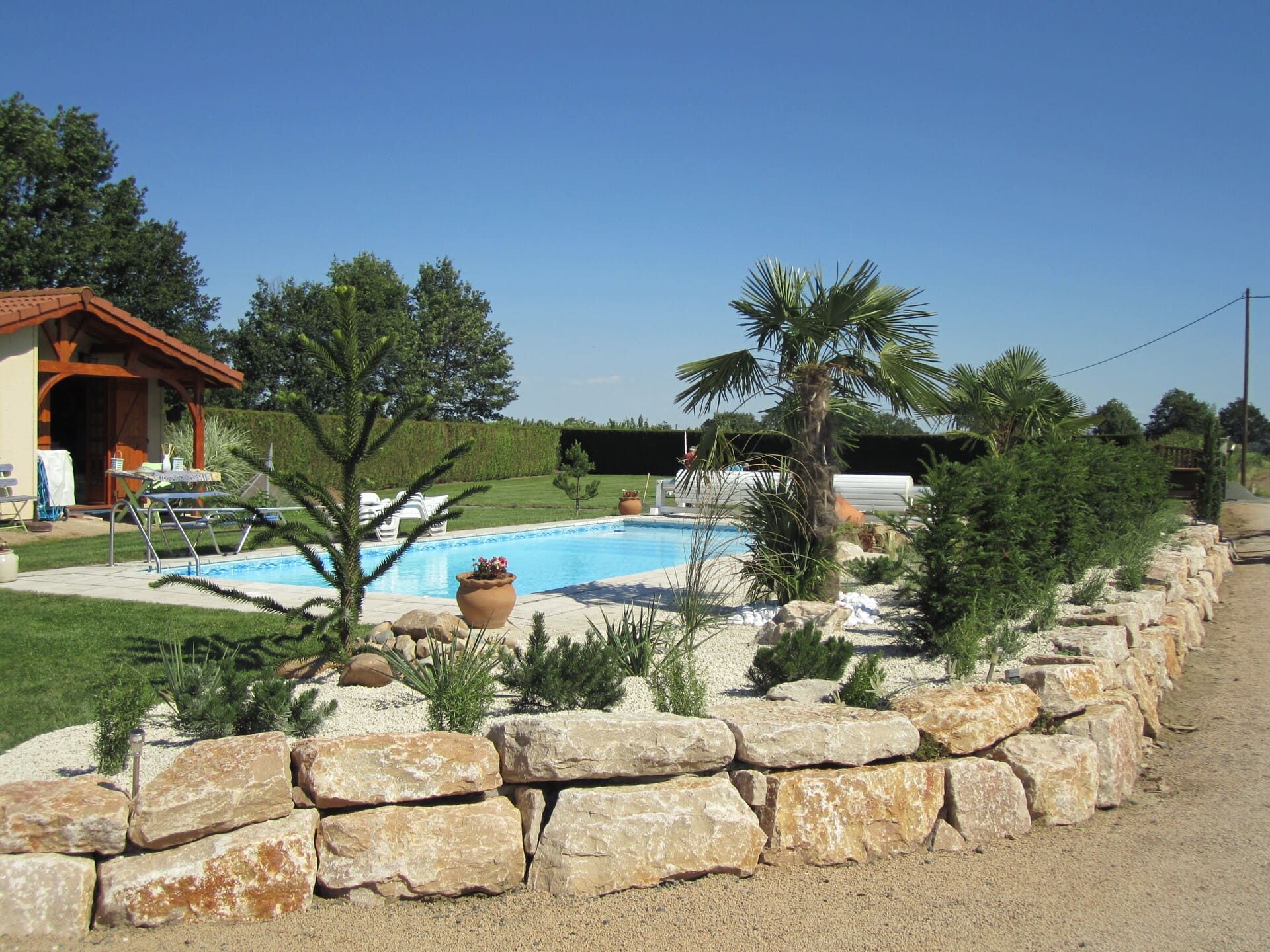 Jardin paysager avec une piscine, entouré de murs en pierre et de diverses plantes, y compris des palmiers et des arbustes, sous un ciel bleu.
