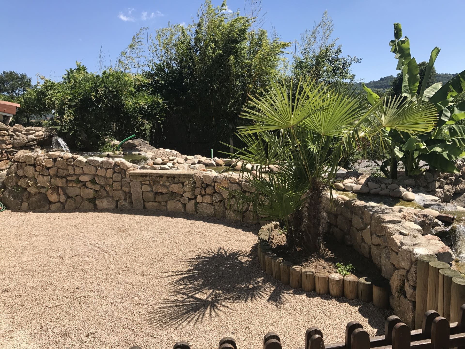 Jardin entouré de murs en pierre, avec des palmiers et des plantes tropicales, et un espace de détente avec des chaises longues.