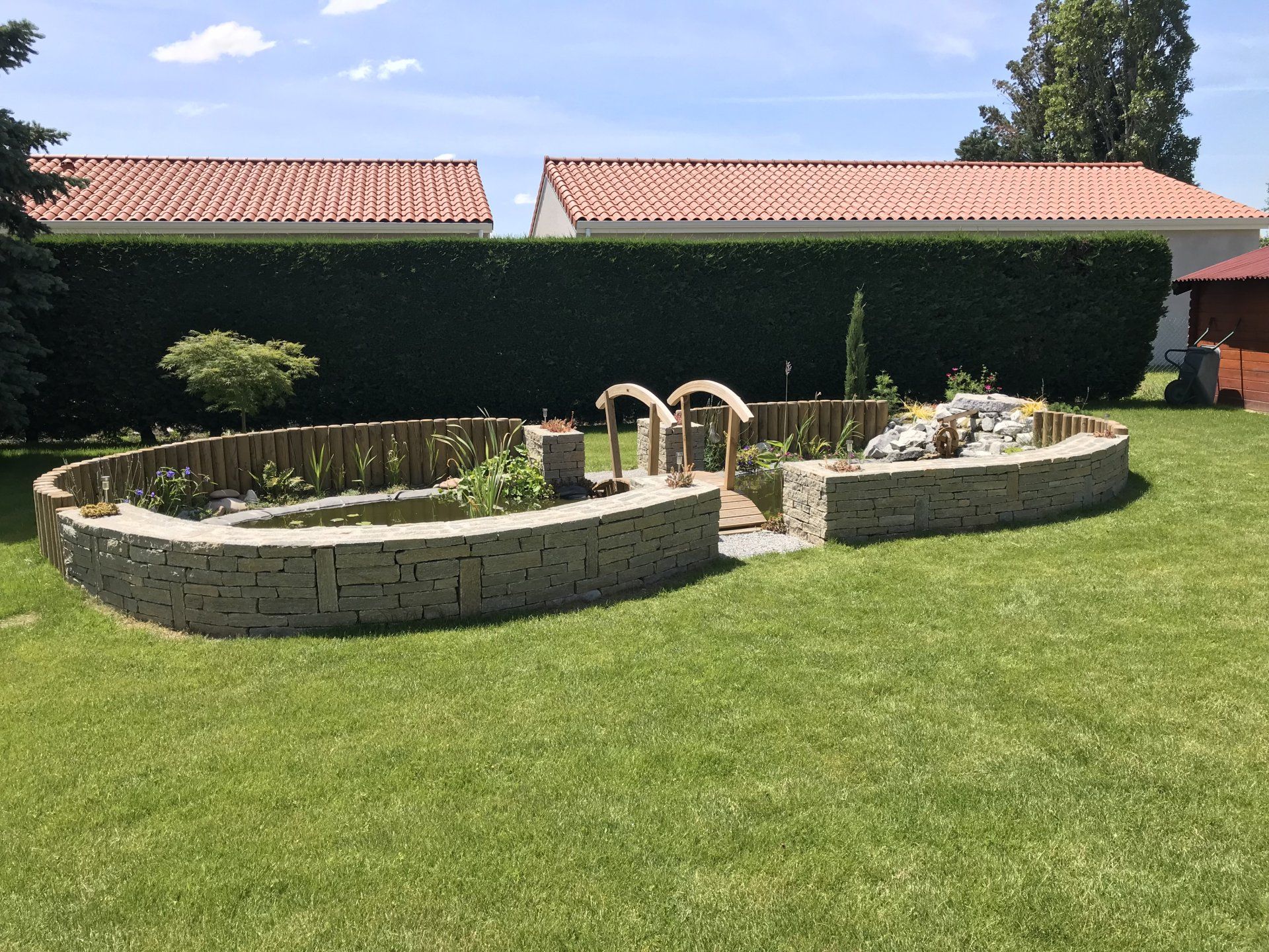 Bassin terminé avec des murs de pierre, un pont en bois, et des plantes aquatiques, entouré d'une pelouse verte et de haies.