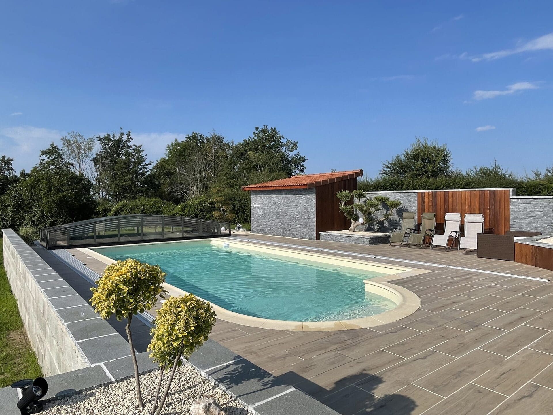 Piscine rectangulaire avec forme arrondie et terrasse en carrelage, chaises longues, et mur de pierre décoratif.