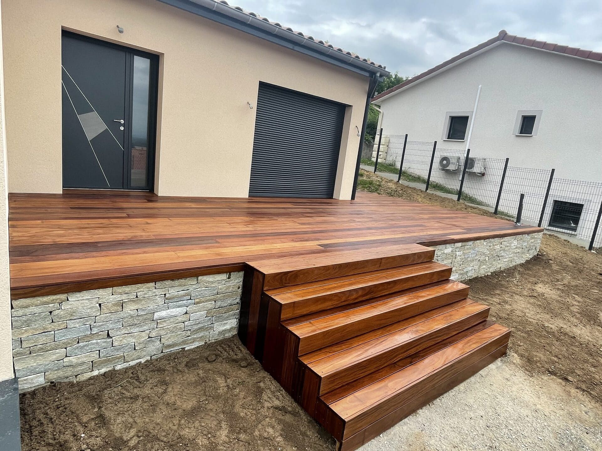 Terrasse en bois avec escalier intégré, devant une maison avec porte moderne et volet roulant.