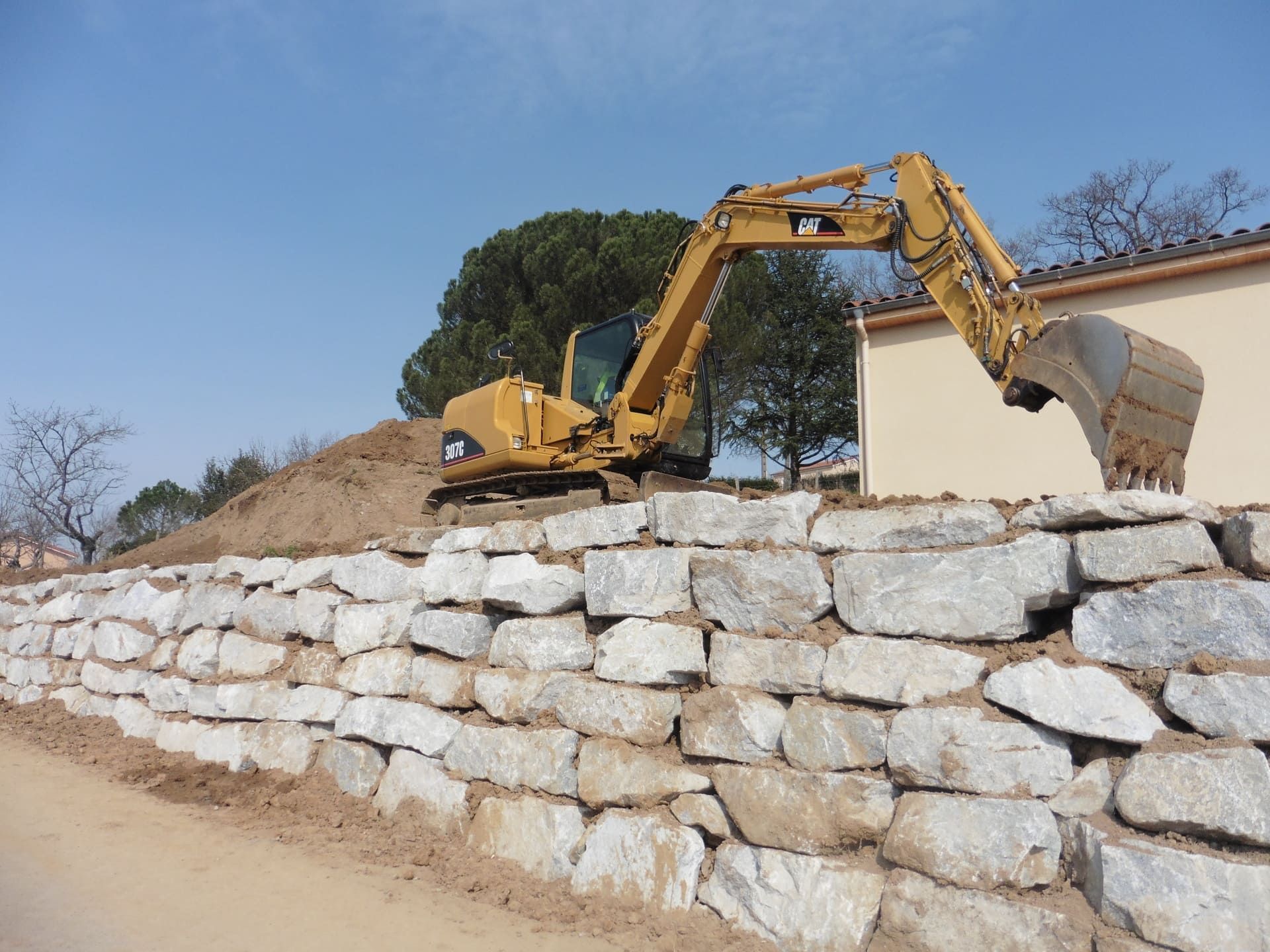 Pelle mécanique hydraulique Caterpillar en train de déplacer de la terre sur un mur de soutènement en pierres grises, avec un bâtiment beige en arrière-plan.