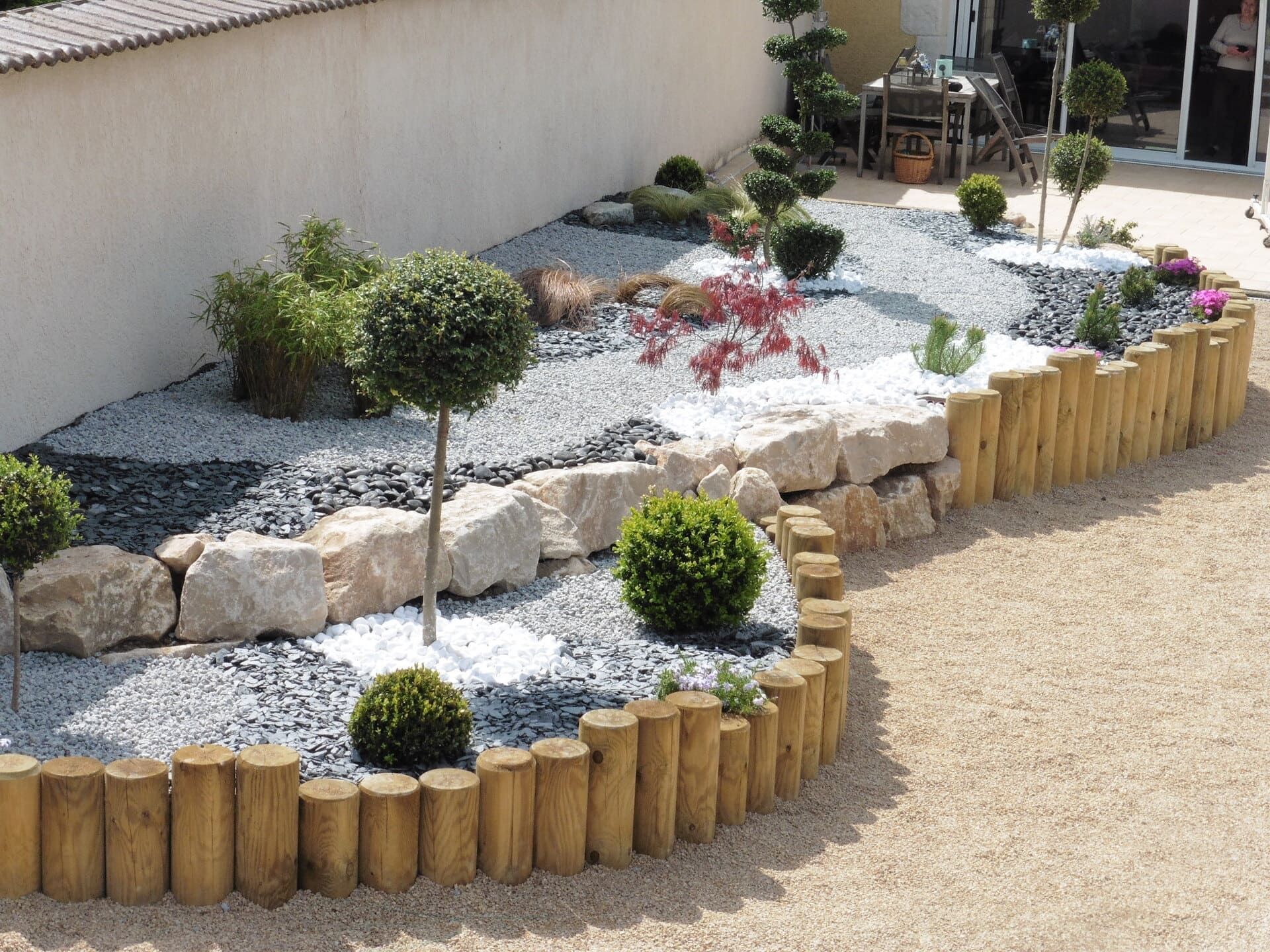 Allée dans un jardin aménagé avec des arbustes et des cailloux décoratifs aux sols et une piscine couverte en arrière-plan.