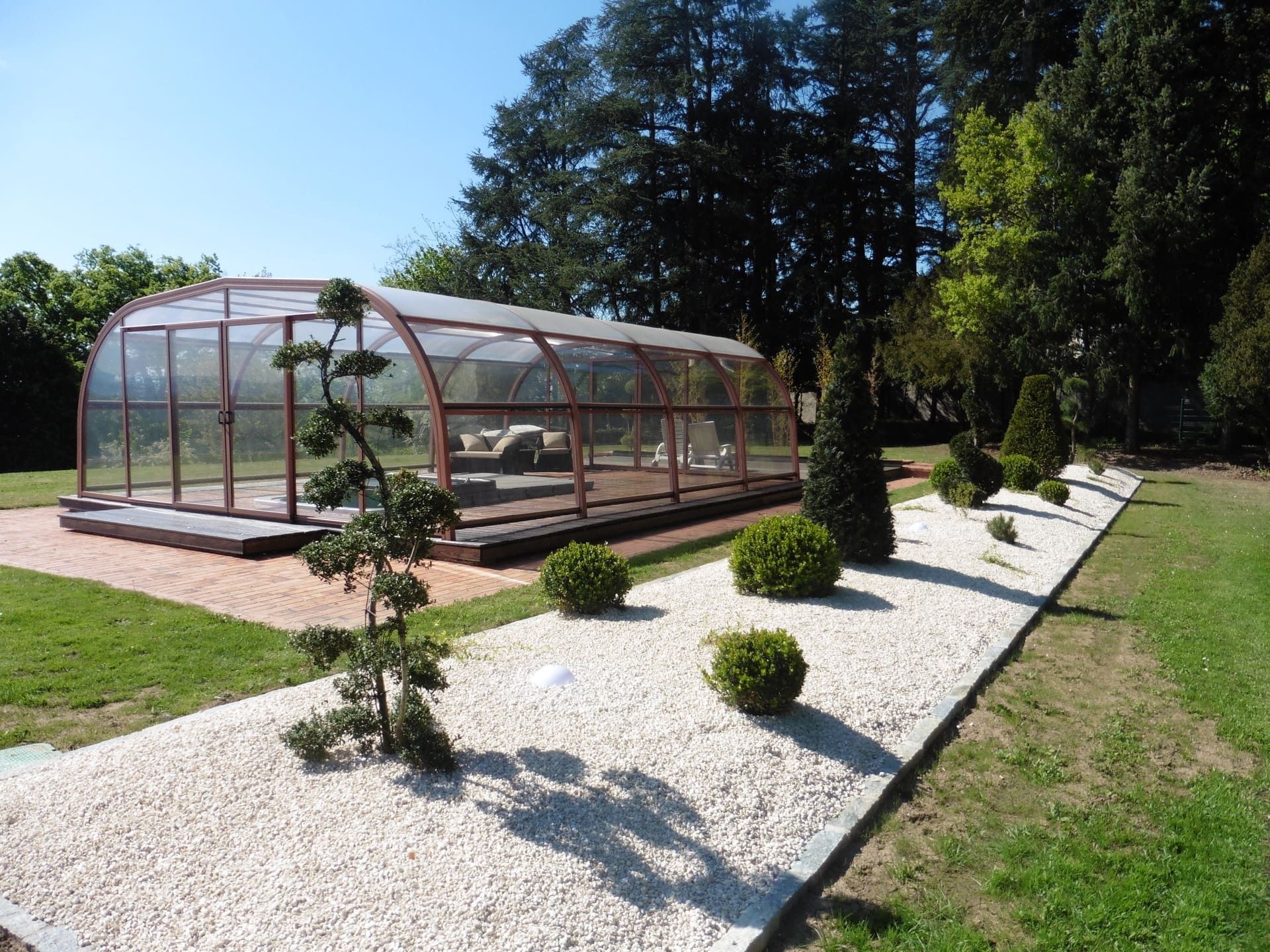 Allée dans un jardin aménagé avec des arbustes et des cailloux décoratifs aux sols et une piscine couverte en arrière-plan.