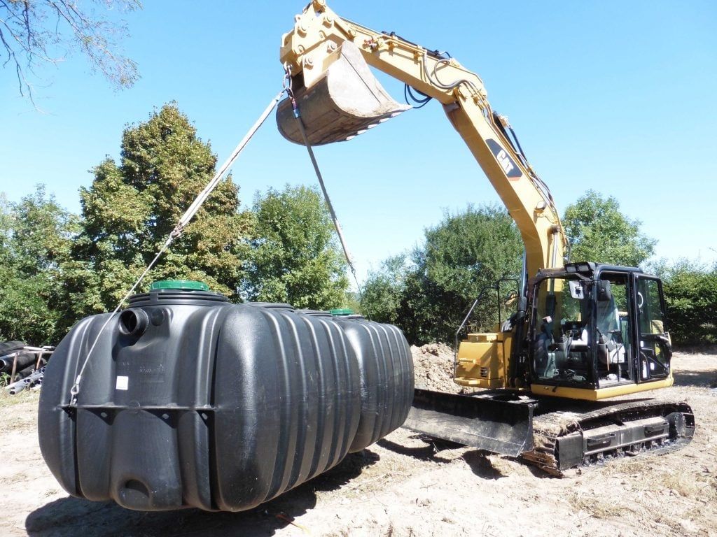Pelleteuse soulevant un réservoir en plastique noir près d'un bâtiment en construction.