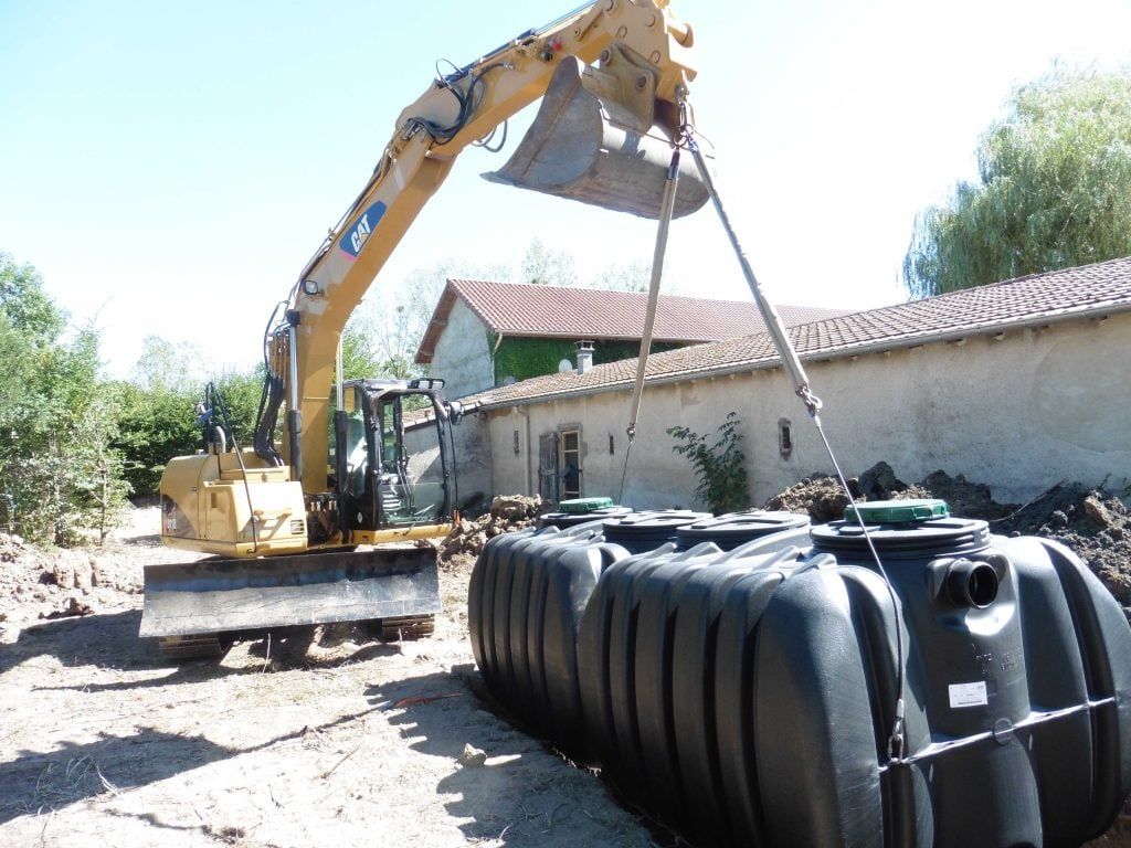 Excavatrice soulevant un grand réservoir en plastique noir avec des sangles.