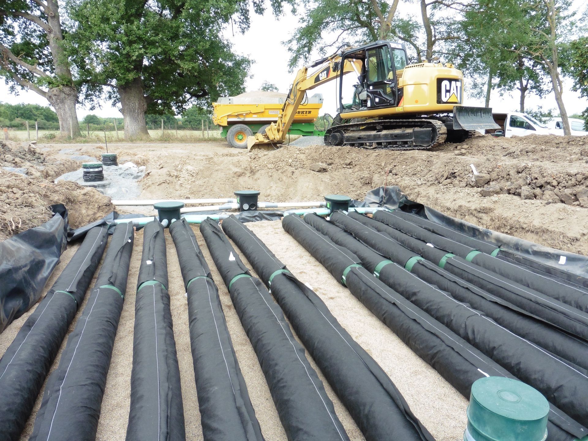 Pelles mécaniques Caterpillar en action sur un chantier de construction, avec des tuyaux de drainage recouverts de géotextile en arrière-plan.