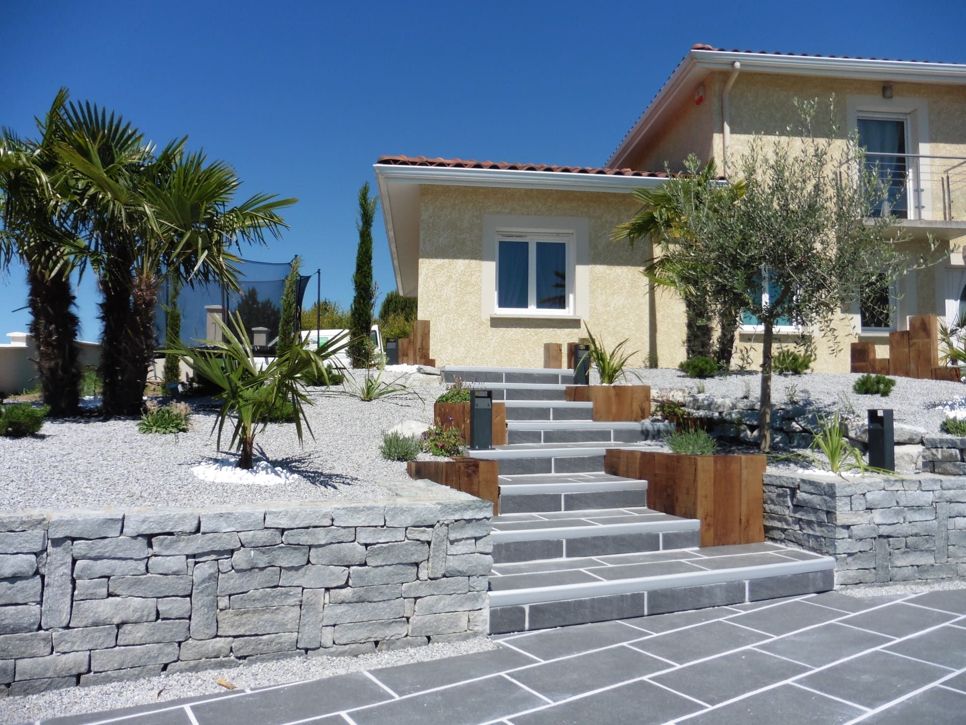 Jardin paysager avec des marches en pierre, des palmiers, et une maison moderne en arrière-plan.