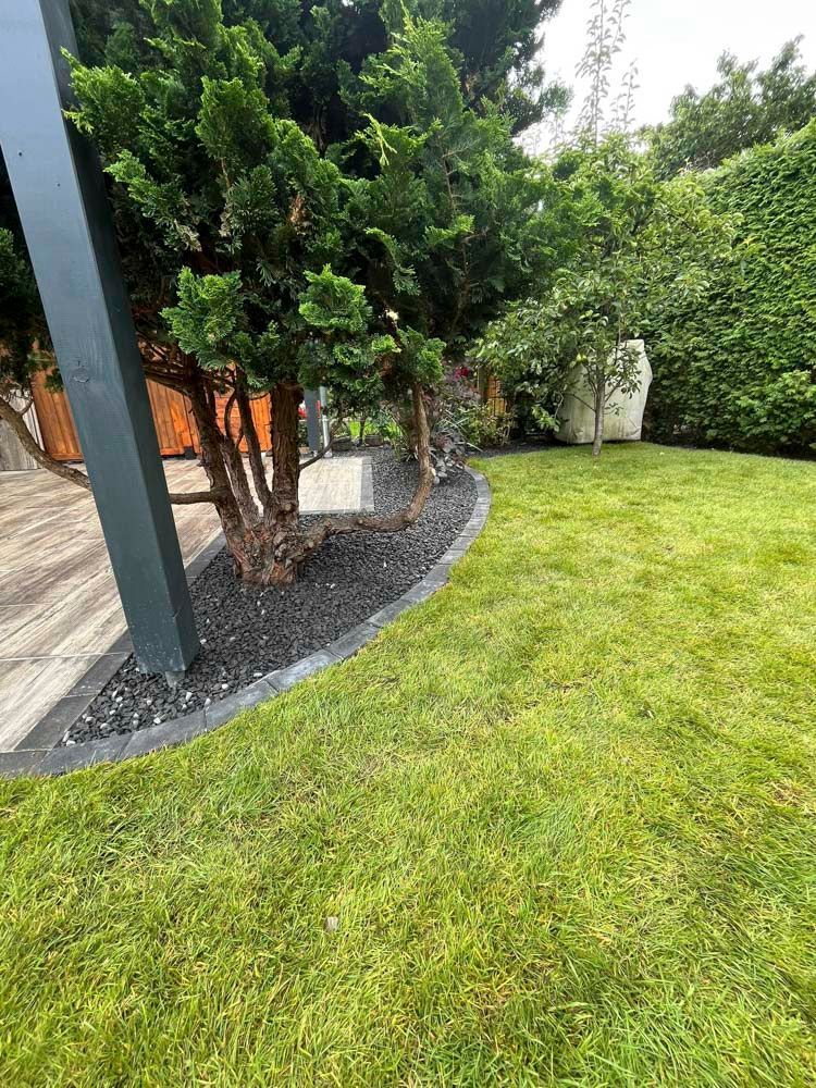 Saftig grüner Rasen mit schwarzem Kiesbett, das an einen Baum und einen dunklen Pfosten in einer Gartenumgebung grenzt.