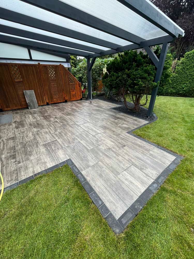 Terrasse mit grauen Fliesen in Holzoptik, eingefasst von schwarzen Pflastersteinen, unter einer grauen Pergola; ein Rasen umgibt die Terrasse.