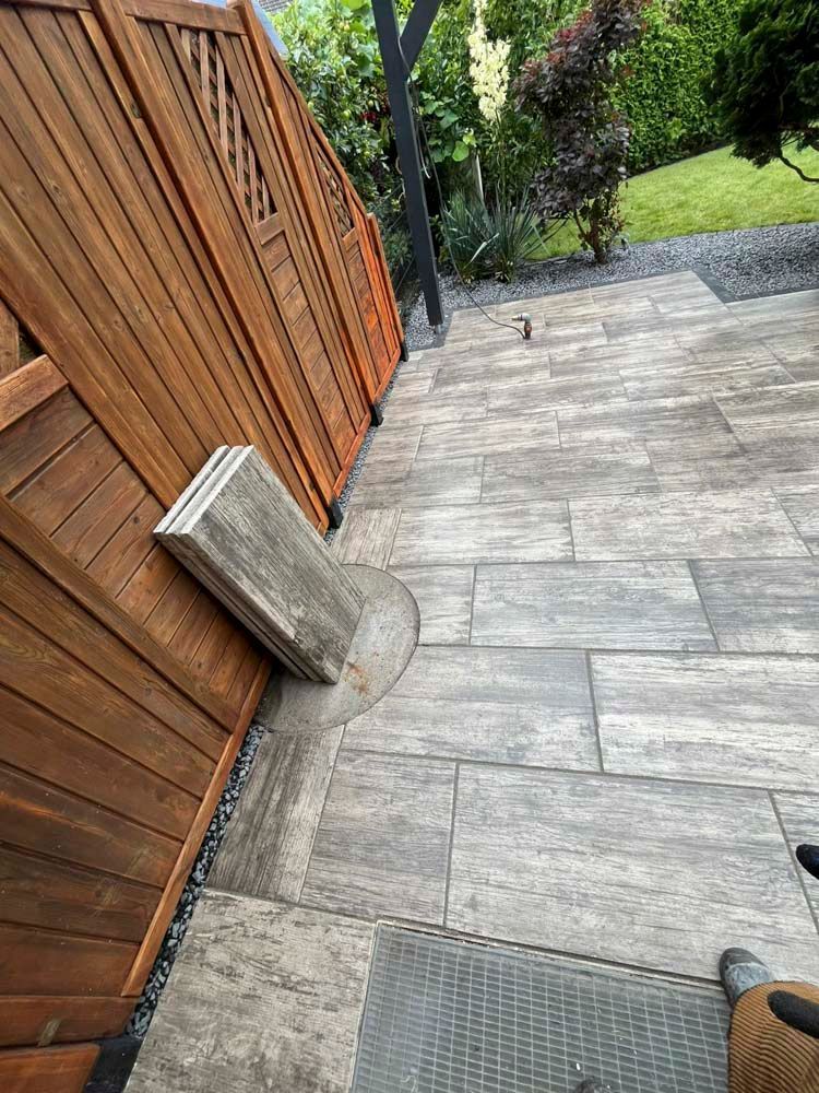 Eine Terrasse im Hinterhof mit grauen Steinpflastern. Links ist ein Holzzaun zu sehen, an dem ein Stapel grauer Pflastersteine lehnt.