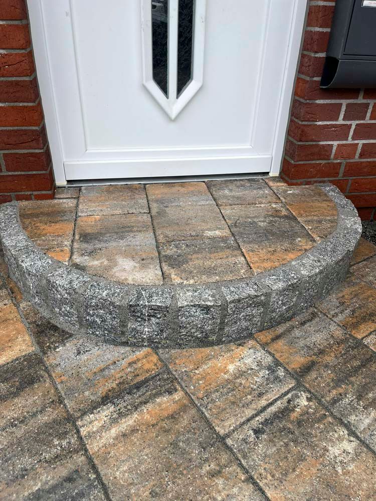 Ein Eingang mit einer geschwungenen Stufe aus grauen Blöcken, die auf mehrfarbigen Pflastersteinen steht. Die Tür ist weiß.