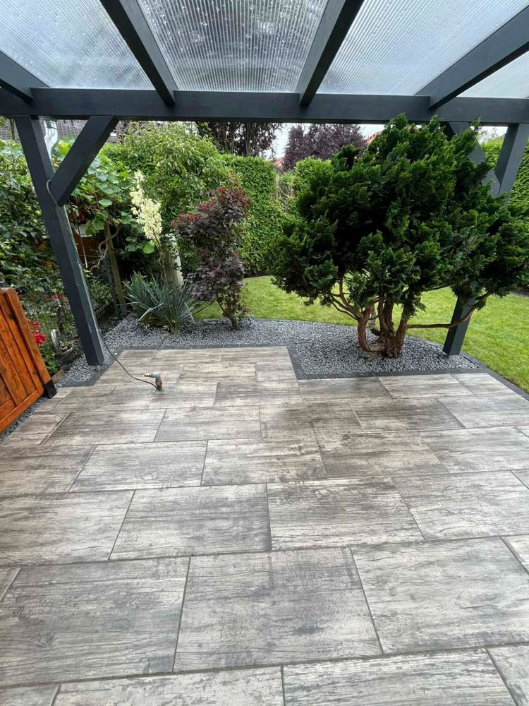 Terrasse mit grauen Steinfliesen, einer Pergola und einem Garten mit Grünflächen und einem kleinen Baum.