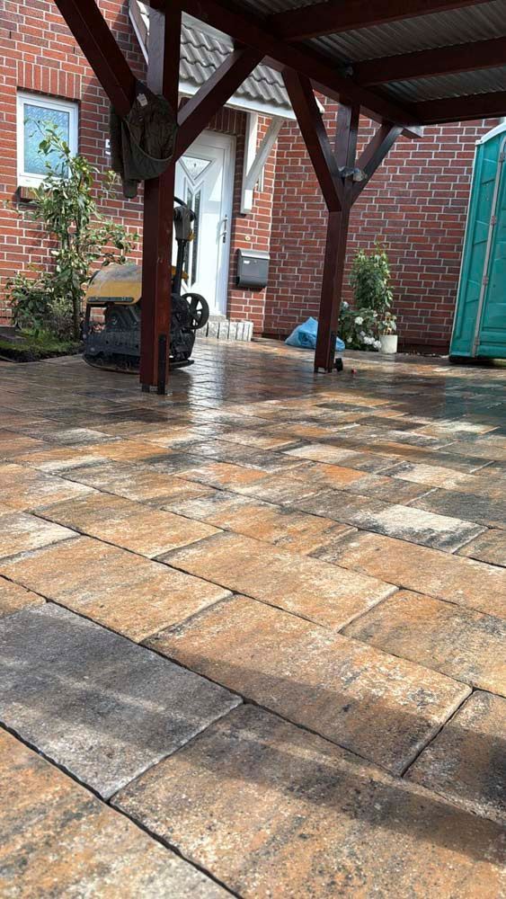 Nasse Ziegelterrasse unter einer Holzkonstruktion, die zum Eingang eines Ziegelgebäudes führt. Eine Verdichtungsmaschine und eine mobile Toilette sind sichtbar.