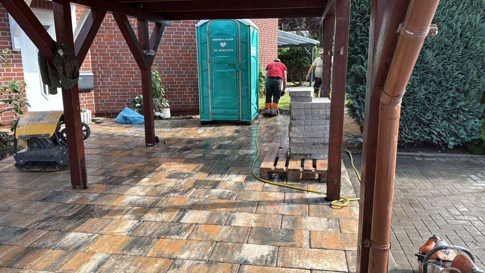 Eine Person in einem roten Hemd und eine weitere Person stehen neben einer grünen mobilen Toilette auf einer gemauerten Terrasse unter einer Holzkonstruktion.