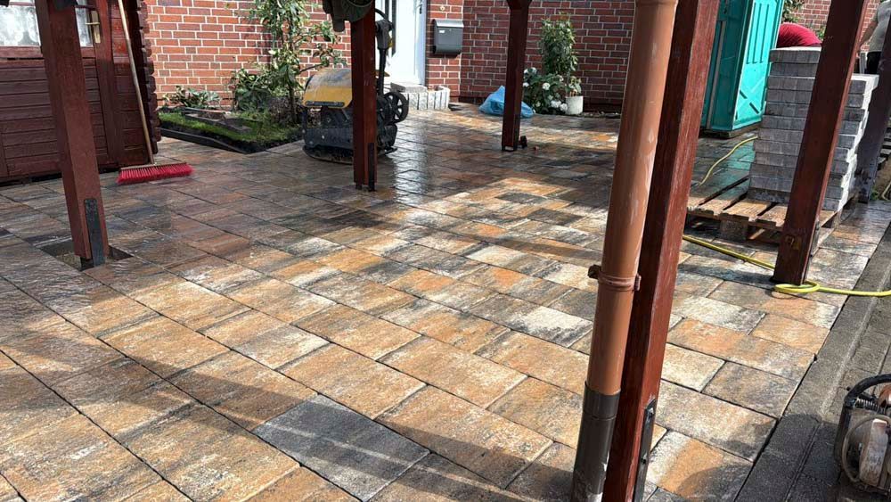 Eine nasse, neu angelegte Terrasse aus hellbraunen und grauen Pflastersteinen unter einer teilweise errichteten Pergola.