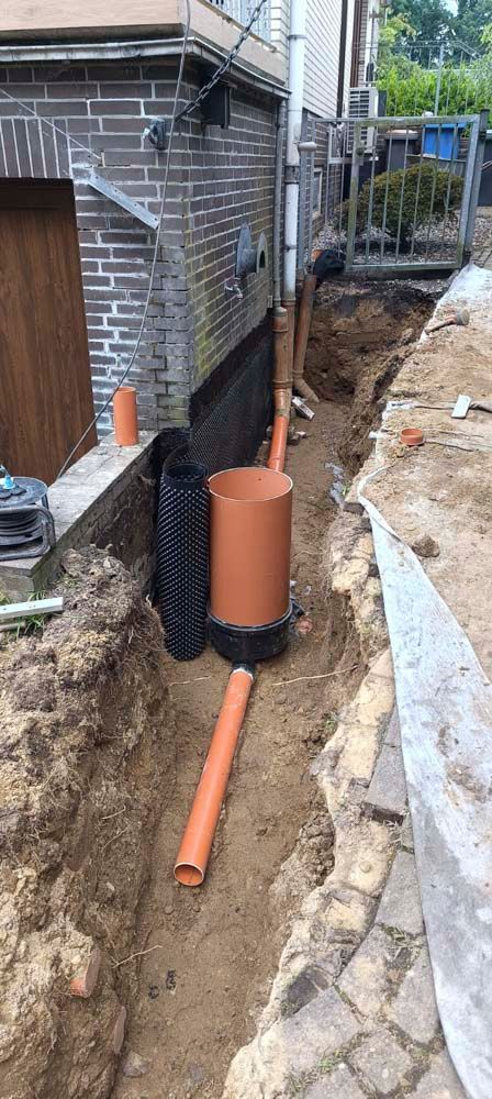 Baustelle mit einem Graben neben einem Backsteingebäude, der Abflussrohre und eine große zylindrische Struktur enthält.