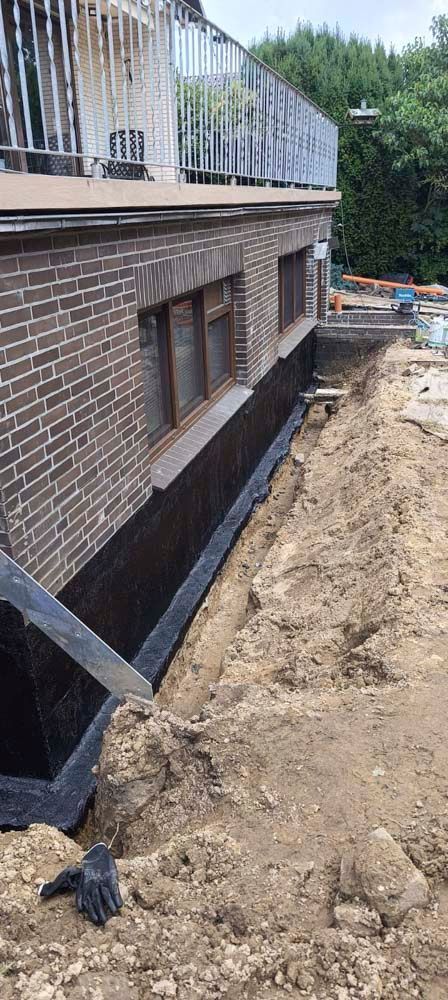 Außenansicht eines im Bau befindlichen Backsteingebäudes mit einem Graben und einer schwarzen Abdichtungsmembran. Ein schwarzer Arbeitshandschuh liegt im Dreck.