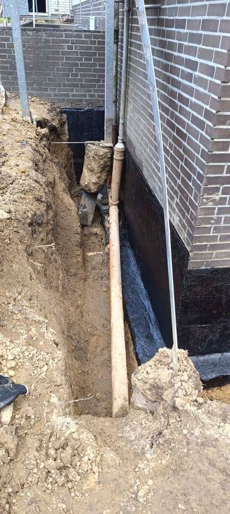 Ein Graben neben einer Ziegelmauer legt ein Rohr frei. Erde und Baumaterialien umgeben den Bereich.