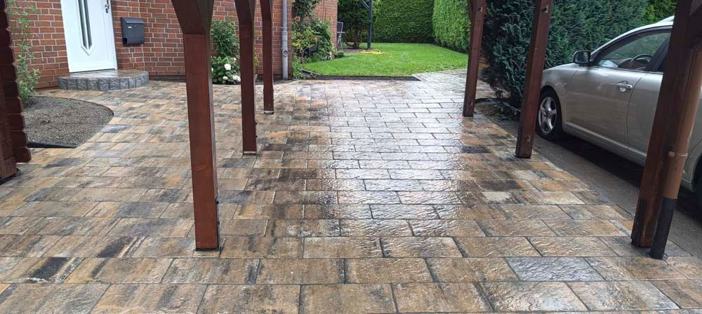 Eine gemauerte Auffahrt mit Holzpfosten stützt einen überdachten Bereich. Rechts parkt ein silbernes Auto, im Hintergrund ist ein Rasen zu sehen.