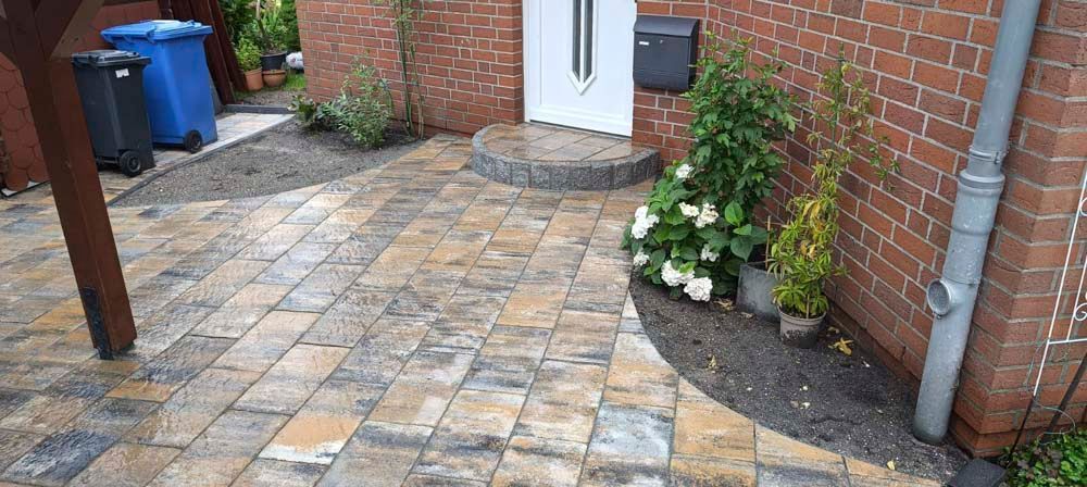 Eine gemauerte Terrasse mit einem Gehweg, der zu einer Haustür, einem Briefkasten und einem Garten mit weißen Blumen führt.