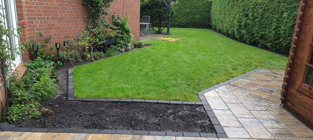 Ein gepflegter Hinterhof mit grünem Rasen, einem Backsteinhaus und einer gepflasterten Fläche. Es gibt ein Blumenbeet mit schwarzem Mulch.