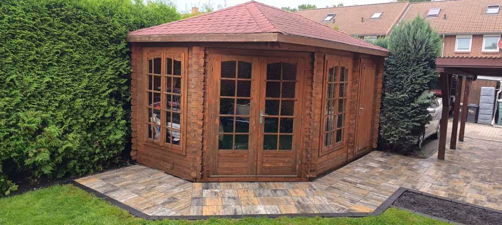 Ein hölzerner Pavillon mit Türen und Fenstern aus Glas ist von einer gepflasterten Terrasse und einem üppigen grünen Rasen umgeben.