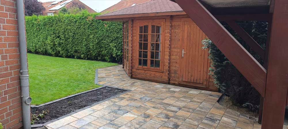 Ein Holzschuppen mit einer gemauerten Terrasse und einem grünen Rasen, eingerahmt von einer hohen Hecke. Die Szene spielt im Freien.