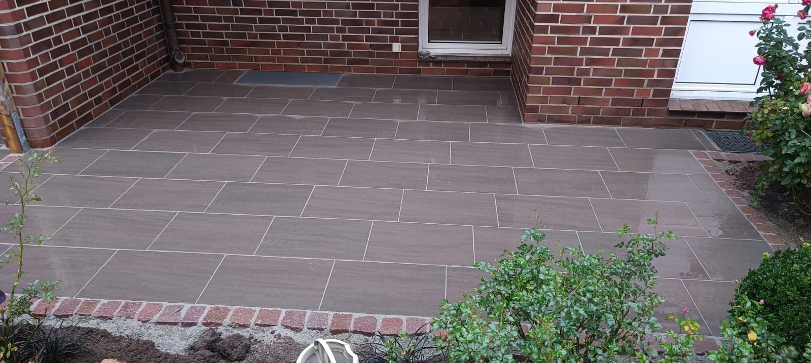 Eine mit grauen Steinfliesen belegte Ziegelterrasse. Ziegelmauern und Grünpflanzen rahmen die Terrasse ein.