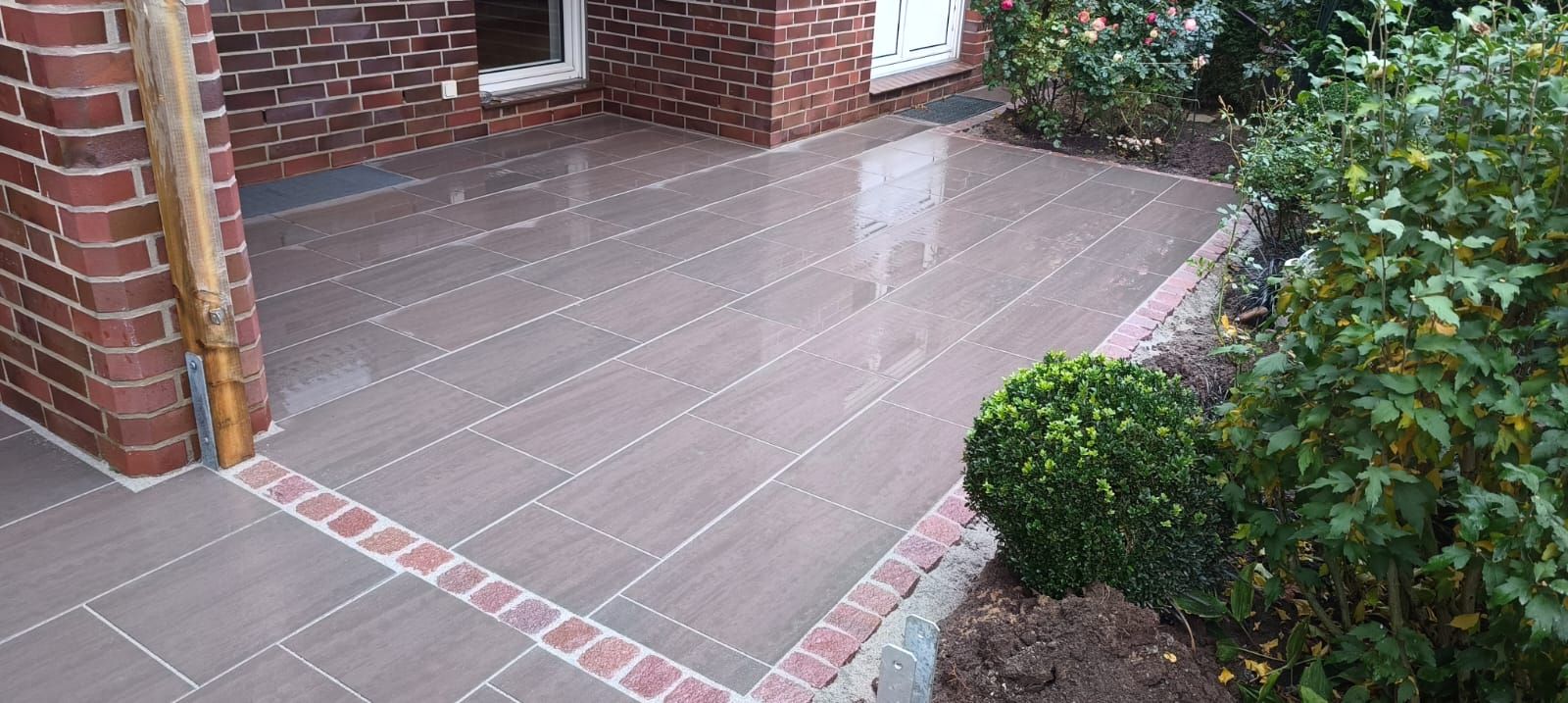 Terrasse mit dunkelgrauen Fliesen und Ziegelumrandung neben einem Backsteingebäude.