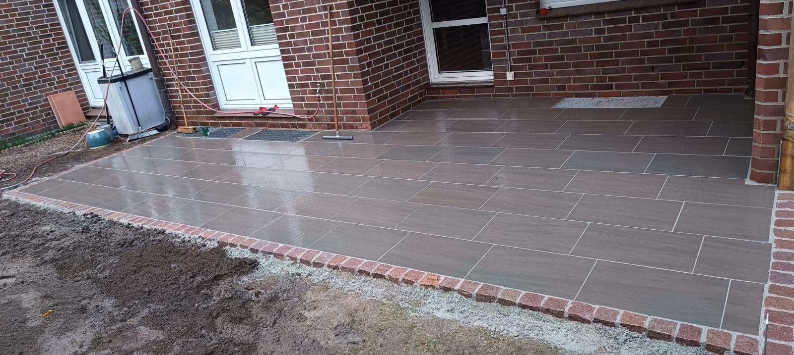 Terrasse aus Ziegelsteinen mit dunkelgrauen Pflastersteinen, roter Ziegelumrandung und rotem Backsteingebäude.
