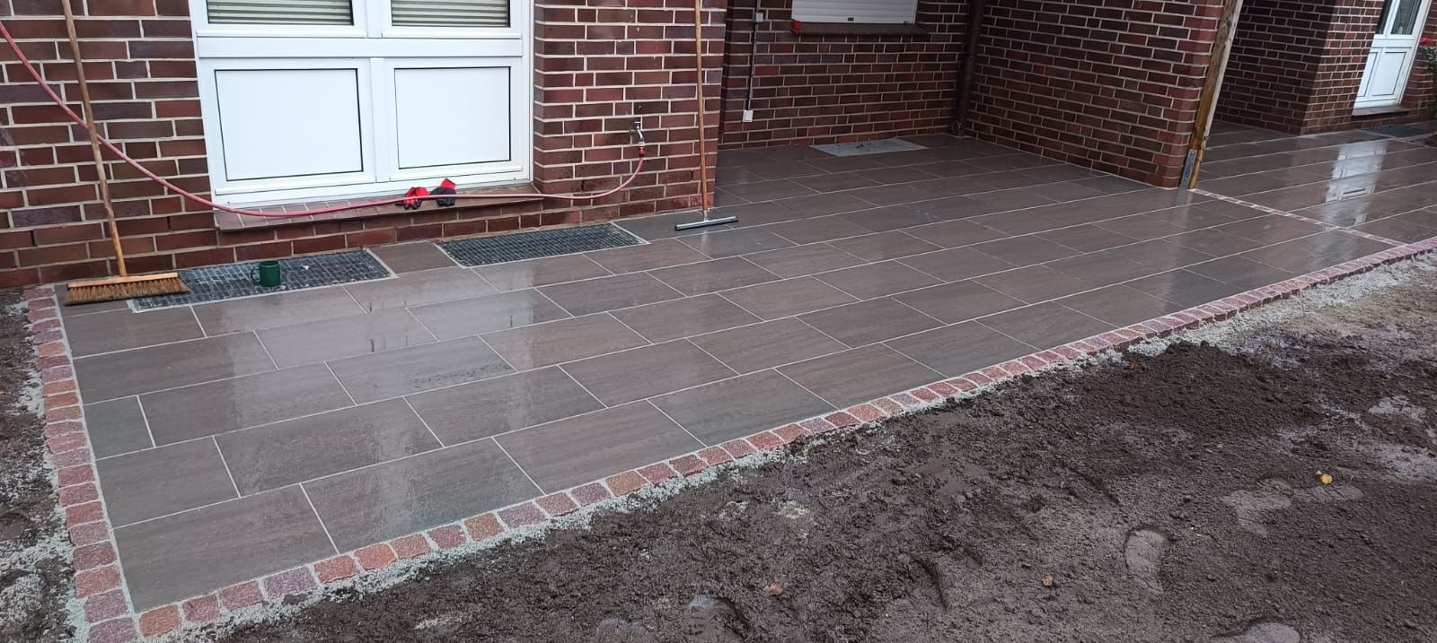 Neu gepflasterte Terrasse mit Ziegelsteinumrandung und nassem Pflaster, neben einem Backsteingebäude.