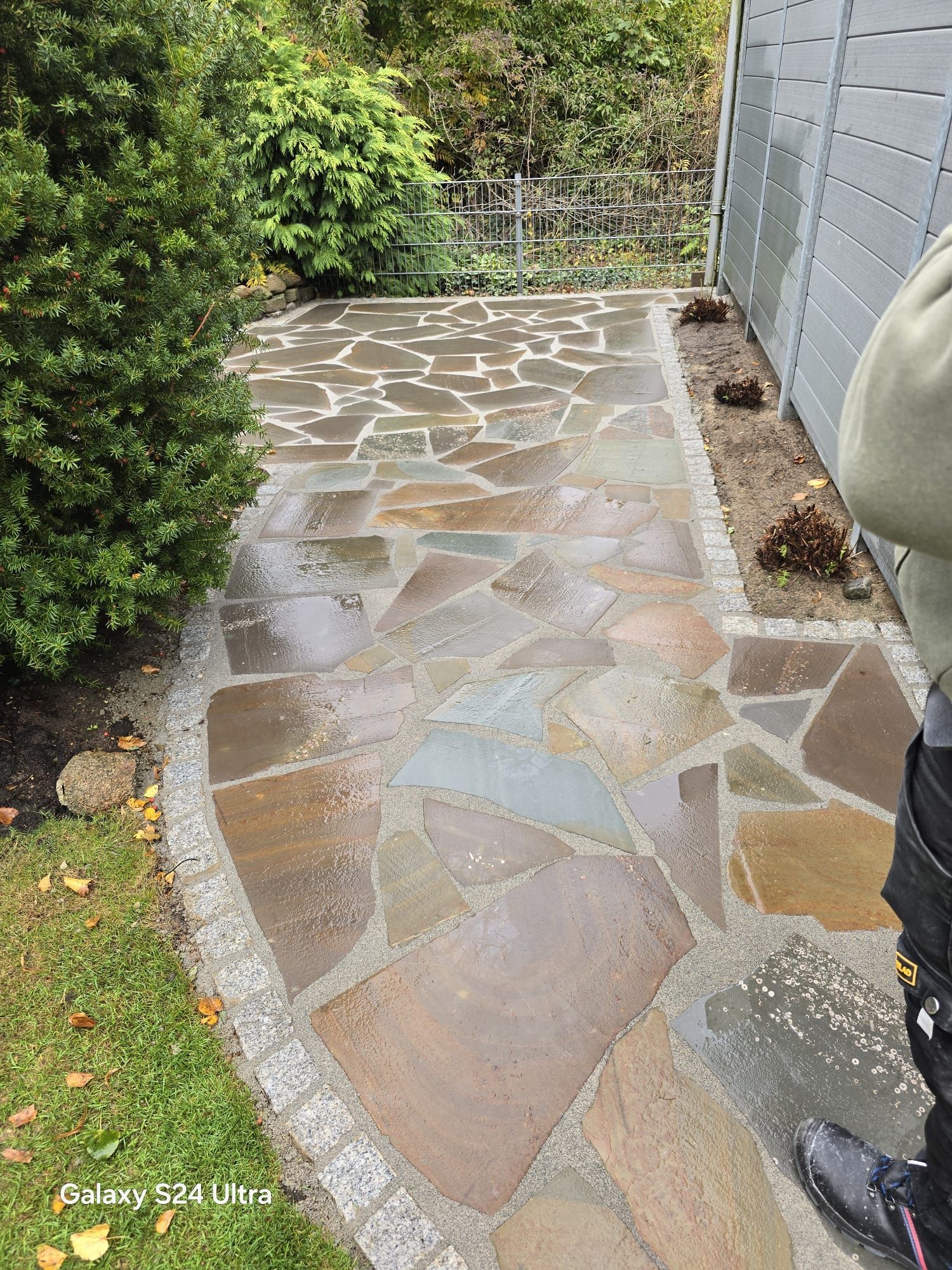 Steinterrasse mit unregelmäßig geformten Fliesen, eingefasst von kleinen Steinen und Gras, neben einem Zaun und Grünflächen.