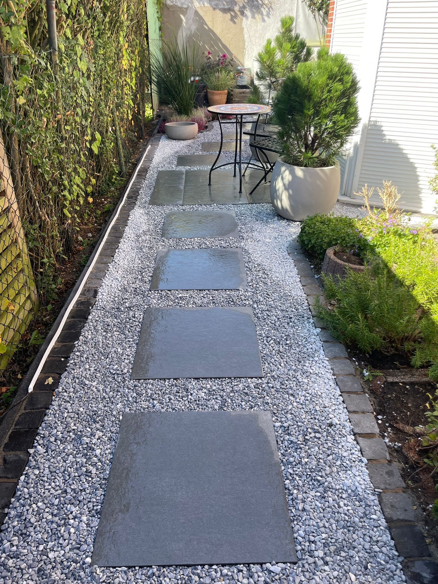 Steinweg durch einen Garten mit Trittsteinen, Kies und Topfpflanzen.