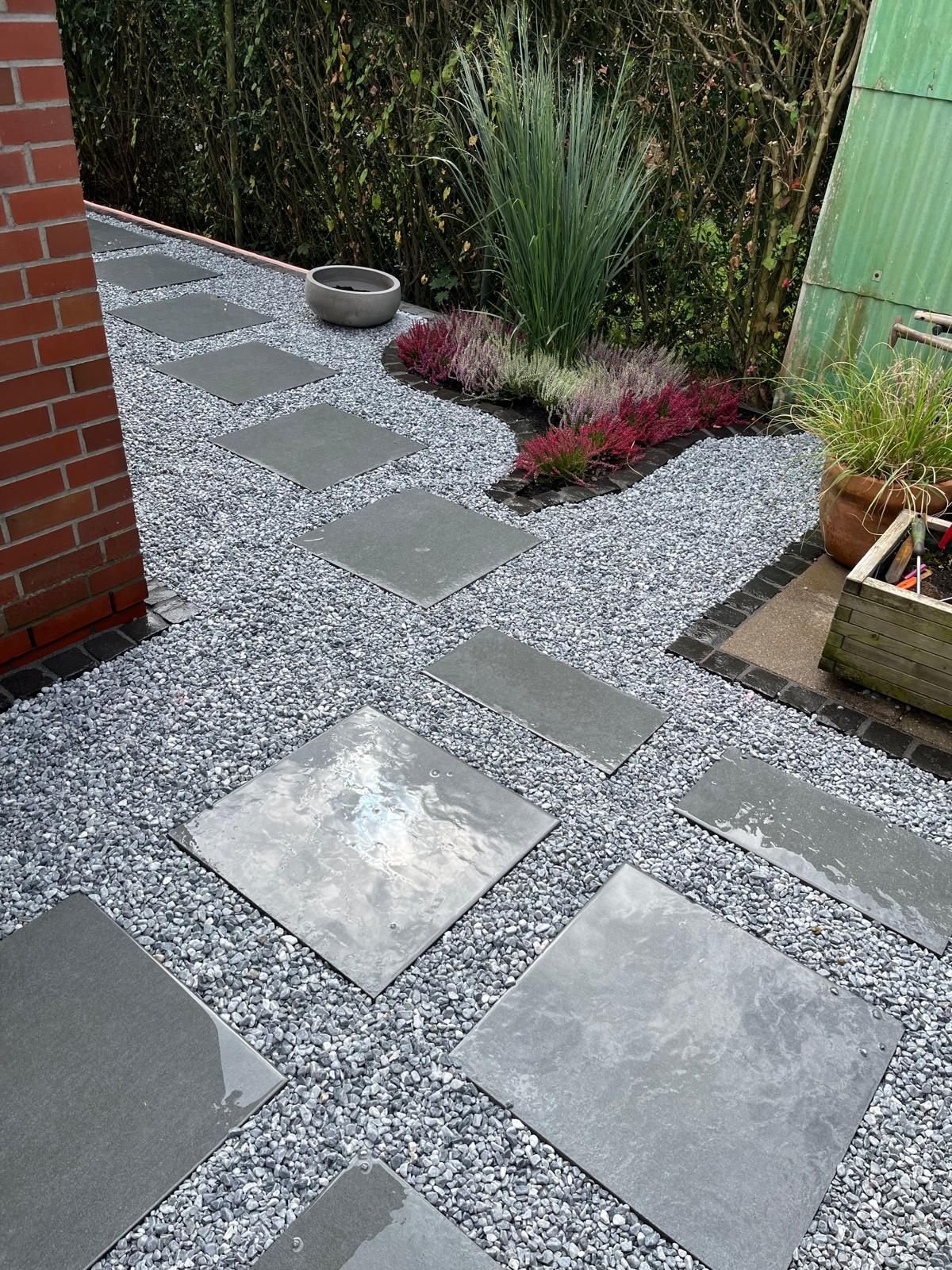 Ein Steinweg mit quadratischen Pflastersteinen, umgeben von grauem Kies, führt zu einem Gartenbeet.