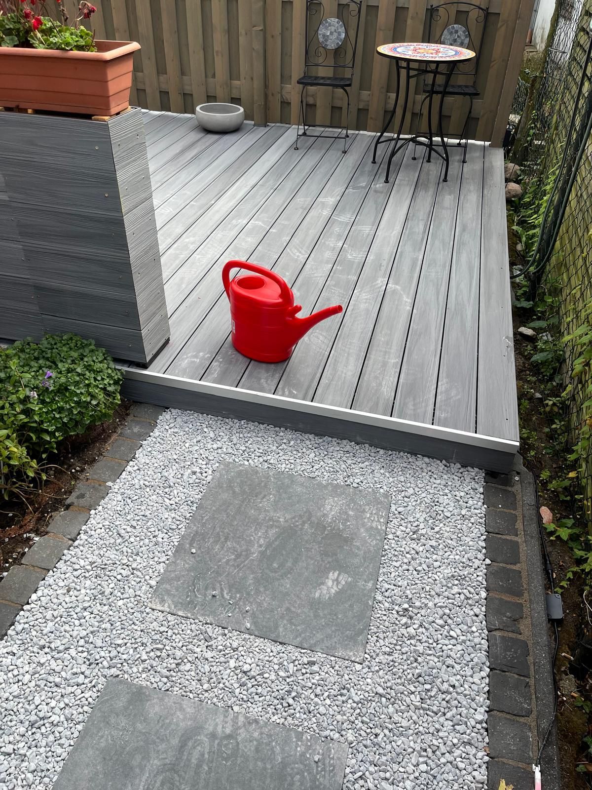 Graue Verbundstoffterrasse mit roter Gießkanne, Trittsteinen und Kiesweg.