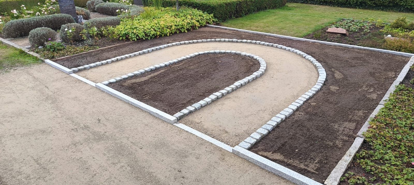 Ein Gartenbeet mit einem geschwungenen Kiesweg und dunkler Erde, umgeben von einer Einfassung und Pflanzen.