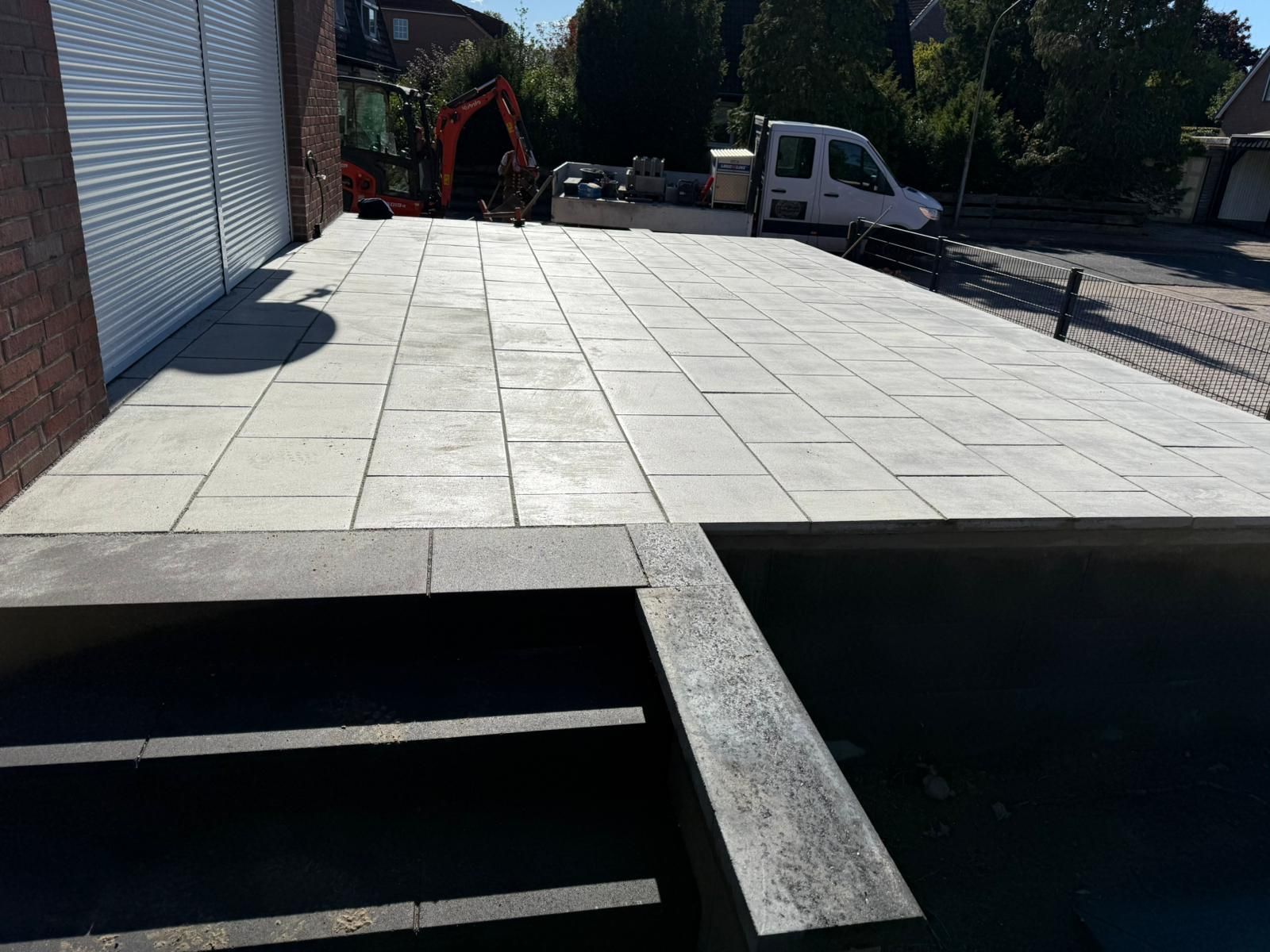 Neu gepflasterte Terrasse mit grauen rechteckigen Fliesen, die zu einer Treppe führt; Baumaschinen sind sichtbar.