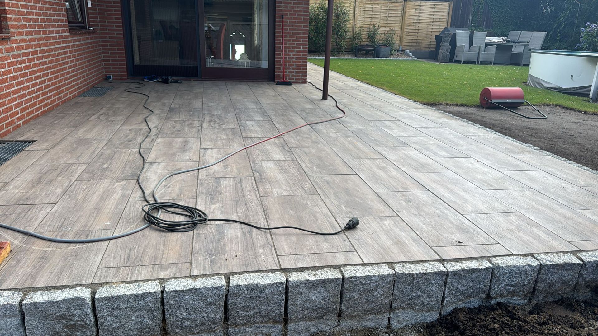 Eine Holzterrasse mit Granitumrandung liegt neben einem Backsteingebäude, im Hintergrund erstreckt sich eine Rasenfläche. Auf der Terrasse verläuft ein Kabel.