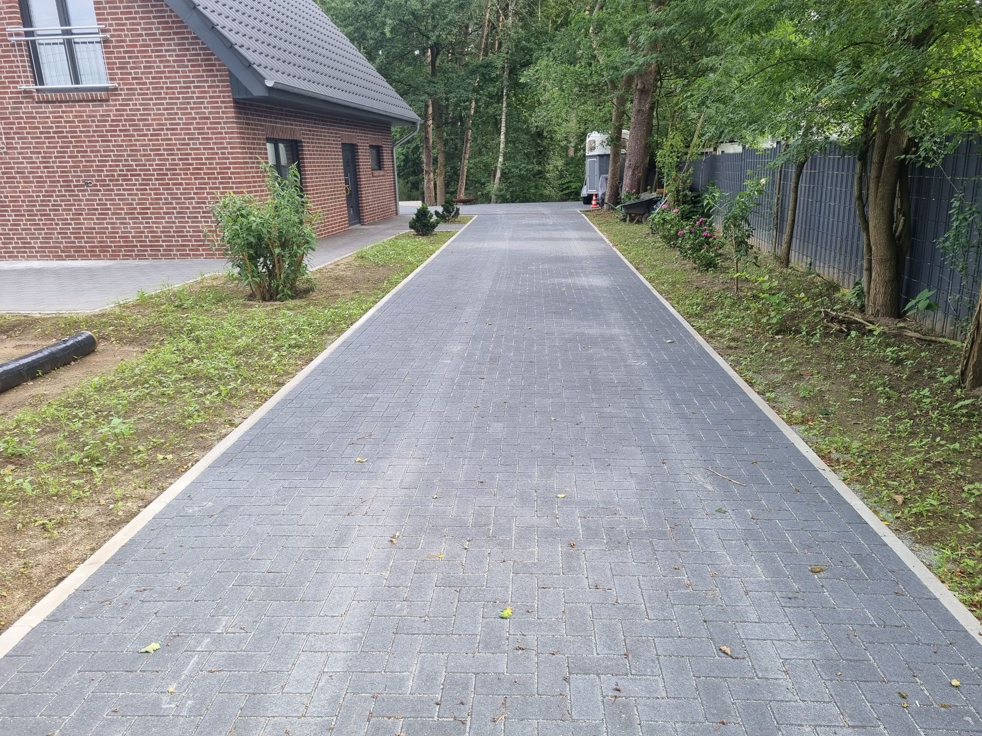 Eine mit Ziegeln gepflasterte Auffahrt führt zu Bäumen, die links von einer Rasenfläche und einem Backsteingebäude begrenzt wird.